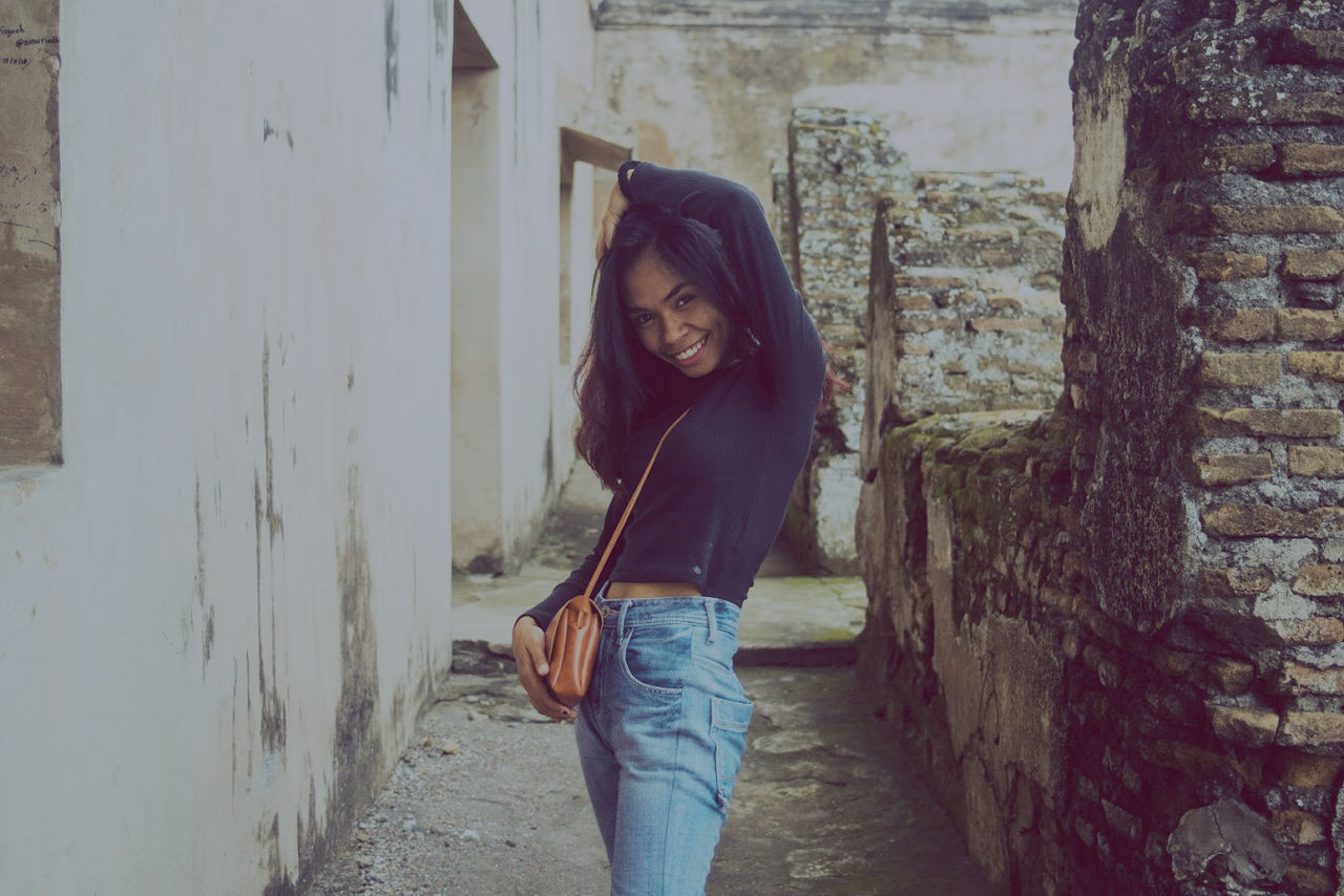 PORTRAIT OF YOUNG WOMAN STANDING AGAINST WALL