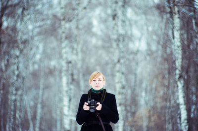 Portrait of young woman holding camera against trees in forest