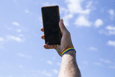 Hand up using the mobile with a lgtb colored bracelet with the sky in the background.