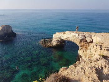 Mid distance view of young woman in bikini standing on mountain by sea