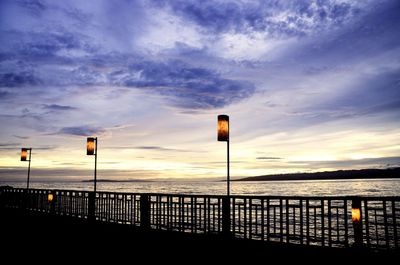 Street lights by sea against sky during sunset