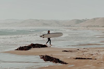 Rear view of woman standing at beach with a surfboard against sky