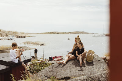 Mother and daughter sitting on rock while brother sitting by with fishing rod during sunny day