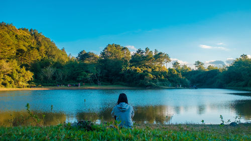 Rear view of young woman sitting at lakeshore against blue sky during sunset