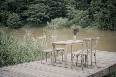 Empty chairs and table arranged on pier by lake against trees