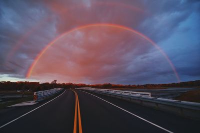 Rainbow over road against cloudy sky during sunset