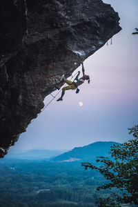 Man clipping quickdraw on overhanging sport climbing route with moon.