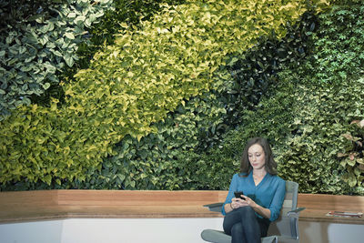 Businesswoman using smart phone in front of green plant wall