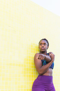 Fit black female athlete in sportswear doing stretching exercises while warming up before training against yellow tiled wall