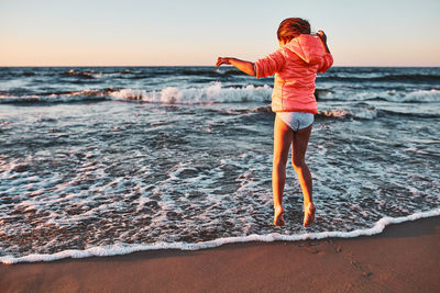 Playful little girl jumping over sea waves on a sand beach at sunset during summer vacation