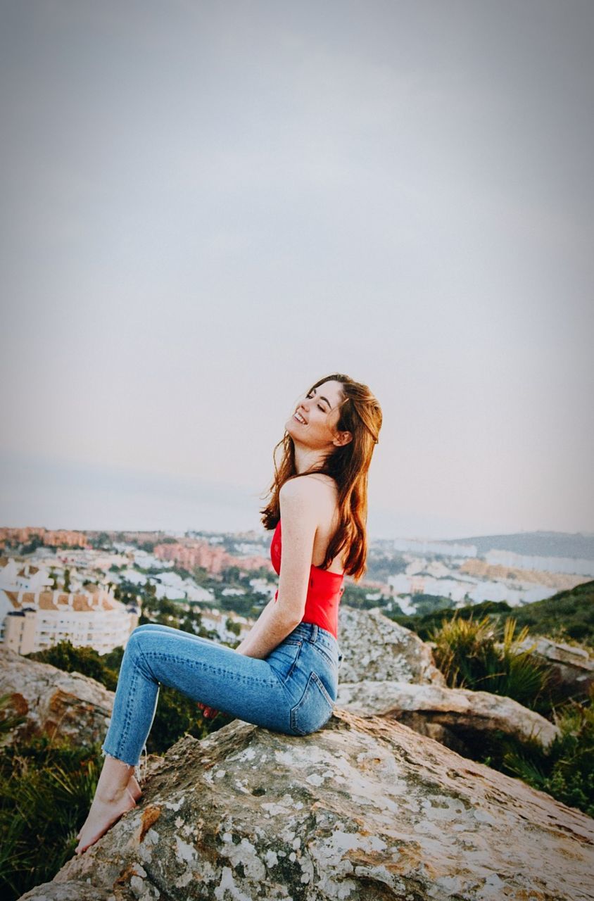 sky, young adult, sea, leisure activity, young women, lifestyles, one person, casual clothing, full length, water, real people, long hair, sitting, beauty in nature, rock, nature, rock - object, hair, beautiful woman, hairstyle, horizon over water, outdoors, freedom
