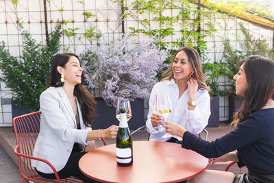 Cheerful adult girlfriends in smart casual clothes sitting at restaurant table and clinking glasses of white wine during meeting on terrace