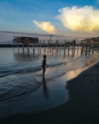 Boy standing on shore at beach against sky during sunset