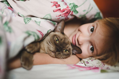 Portrait of cute girl lying with burmese cat under the blanket at home