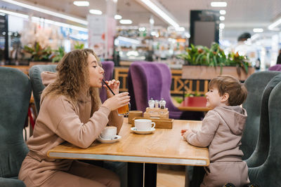 Young curly haired mother and her little son are having breakfast or lunch in a cafe, sitting 