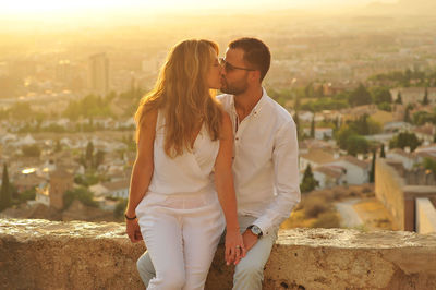Young romantic couple kissing while sitting on retaining wall against cityscape