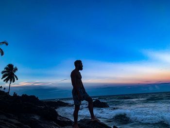 Man standing on rock at beach against sky during sunset