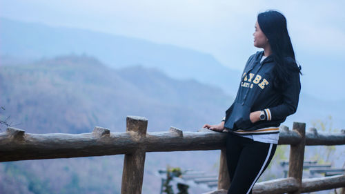 Side view of young woman standing by railing against mountains and sky