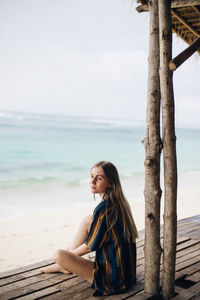 Young woman sitting on shore at beach against sky