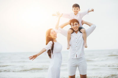 Father carrying son on shoulders by woman at beach against sea and sky