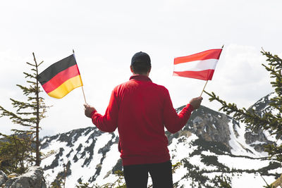 Rear view of mature man holding national flags while standing against snowcapped mountain