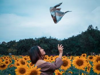 Side view of woman catching scarf while standing on sunflower against sky