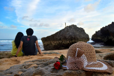Close-up of hat and rose at beach against couple sitting in background