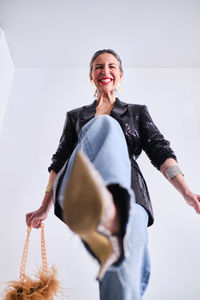 From below of positive female with stylish handbag looking at camera while raising leg in light studio on white background