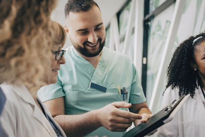 Smiling male healthcare worker discussing with doctor over clipboard in hospital