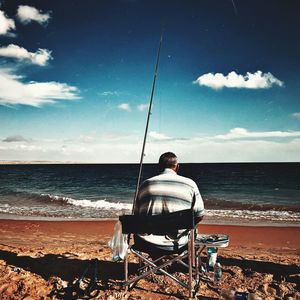 Rear view of man sitting at beach against sky
