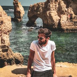 Young man wearing sunglasses while standing against rock formations in sea