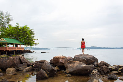 Rear view of woman standing on rock by river against cloudy sky