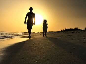 Silhouette father and daughter walking at beach against sky during sunset