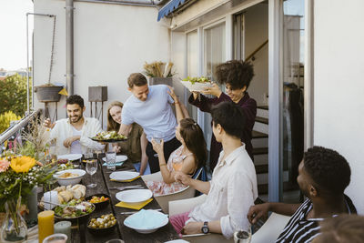 Happy man and woman serving food to group of friends sitting at dining table during dinner party in balcony