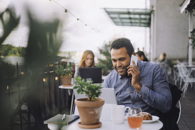 Smiling male entrepreneur talking on smart phone while sitting with laptop at cafe