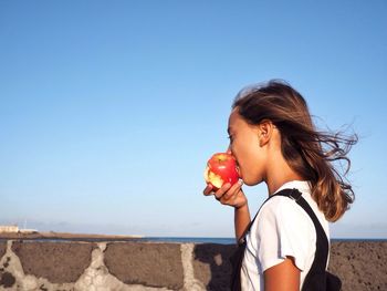 Girl eating apple against clear blue sky