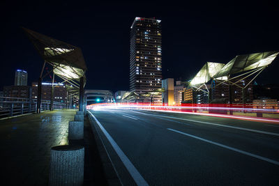 Light trails on road by buildings against sky at night