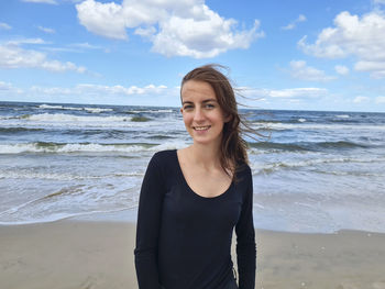 Portrait of young woman standing at beach against sky