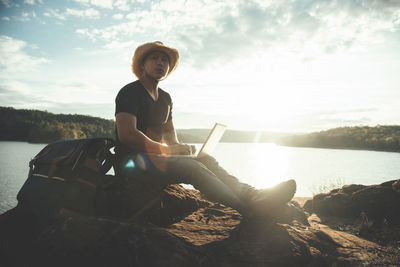 Young woman sitting on rock at shore against sky