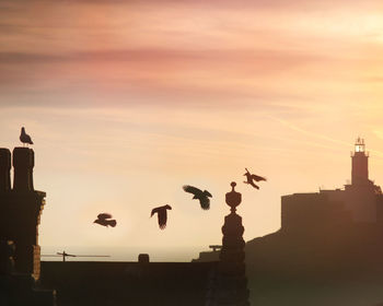 Birds flying by lighthouse against sky during sunset