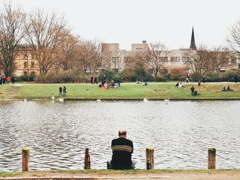 Rear view of man sitting on riverbank by buildings against clear sky