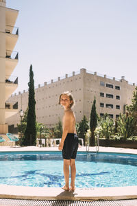 Full length of smiling boy standing by swimming pool against sky