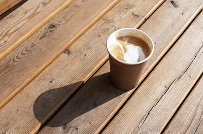 Latte or cappuccino with in beige paper cup on wooden striped table outside, drinking coffee