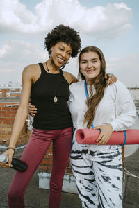Portrait of smiling friends with exercise mat and water bottle standing on rooftop