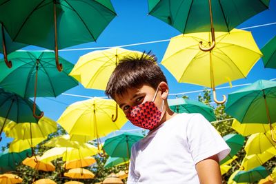 Low angle portrait of boy wearing flu mask standing against umbrellas