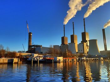 Power station on riverbank emitting smoke against clear blue sky