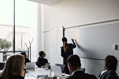 Colleagues looking at businesswoman writing on whiteboard during meeting in board room