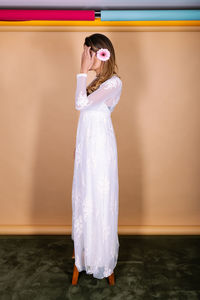 Full body side view of bride in white elegant wedding dress with flower in hair standing in light studio during wedding celebration