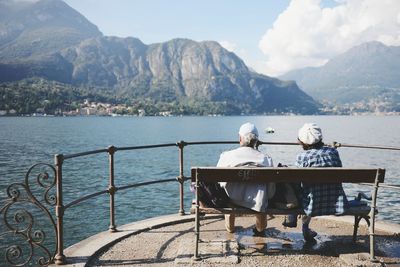Rear view of woman sitting on bench by lake against mountains