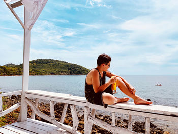 Boy drinking juice in glass while sitting on railing by sea against sky